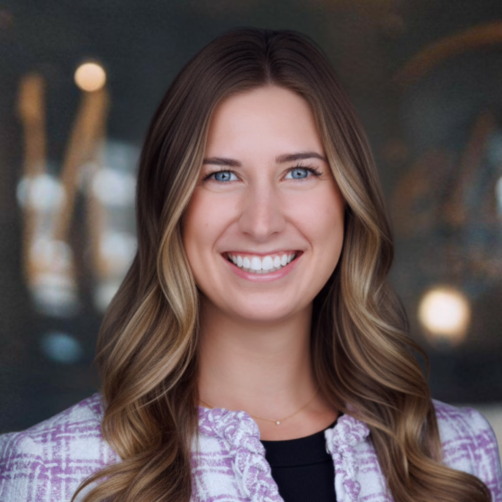 Professional headshot of a smiling woman with long wavy light brown hair, wearing a purple patterned blazer and black top against a neutral blurred background